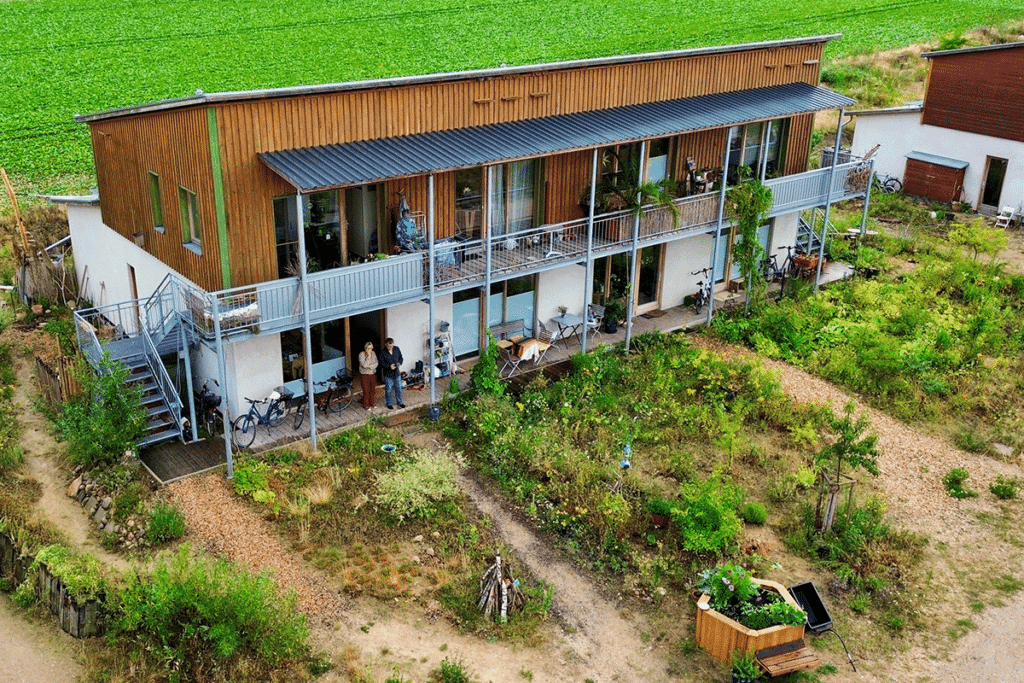 Abbildung: Im Vordergrund sind natürlich angelegte Vorgärten zu sehen. Das Mehrparteienhaus im Hintergrund besteht aus zwei Geschossen, wovon das untere eine helle Fassade und das obere eine Holzfassade hat. Vor dem Haus verlaufen auf beiden Etagen Laubengänge.